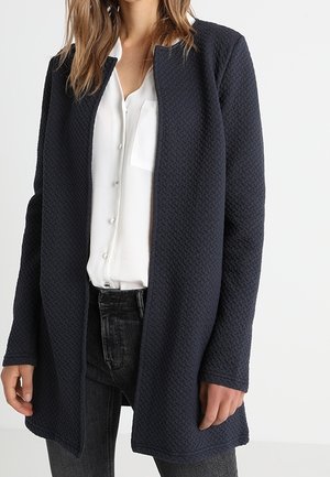 Mujer que lleva una chaqueta larga azul marino texturizada con abertura frontal sobre una blusa blanca abotonada y vaqueros oscuros sobre un fondo liso.