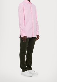 Man i en ljusrosa långärmad skjorta med knappar, mörkgröna byxor och vita snörskor, stående mot en vit bakgrund.