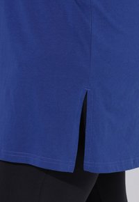 Blaues Baumwolloberteil mit geradem Schnitt, das am Saum seitliche Schlitze aufweist, kombiniert mit schwarzen Leggings. Glatte Textur und schlichtes Design.