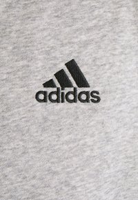 Tela de algodón gris con una textura suave que presenta un logo bordado de Adidas en negro en forma de tres rayas diagonales.