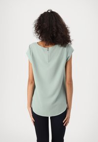 Blouse vert menthe à manches courtes, col rond, détail de fermeture éclair au dos et ourlet incurvé. Texture lisse et coupe décontractée.