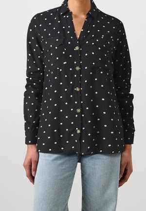Chemise noire à manches longues à boutons avec des pois blancs, poches poitrine, portée avec un jean bleu clair.