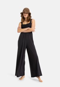 Sort jumpsuit med brede ben og smocket bodice. Båret med en tekstureret brun bucket hat. Modellen står barfodet med armene krydset.