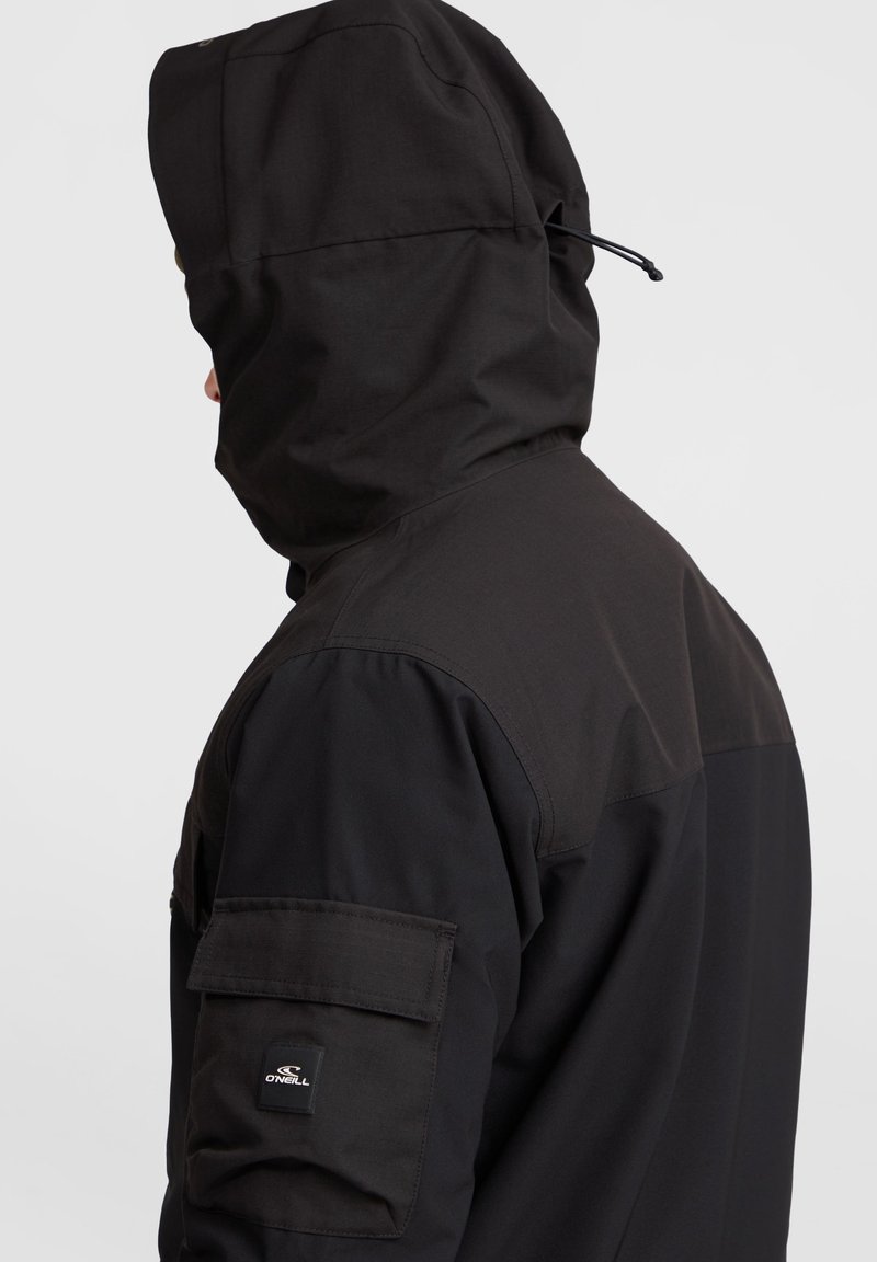 Czarna kurtka z kapturem i wysokim kołnierzem, wykonana z fakturowanej tkaniny, z łatką na rękawie z logo. Regulowany sznurek w kapturze.