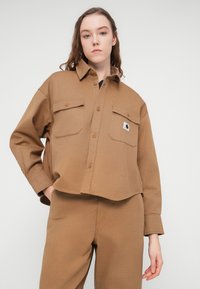 Καφέ κοντομάνικο πουκάμισο από ανθεκτικό ύφασμα με μυτερο κολάρο, κούμπωμα με κουμπιά και δύο τσέπες στο στήθος. Διαθέτει ένθετο με λογότυπο.