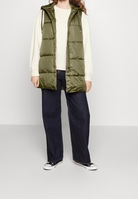 Oliwkowa puchowa kamizelka z kapturem, z dużymi pikowanymi panelami, zamkiem błyskawicznym i bocznymi kieszeniami, noszona na jasnym swetrze i ciemnych dżinsach.