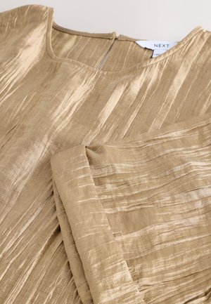 Zlato obarvan zgornji del s teksturo navpičnih prog, zaobljenim izrezom, dolgimi rokavi in zapiranjem na hrbtni strani. Izdelano iz lahkega, zračnega blaga.