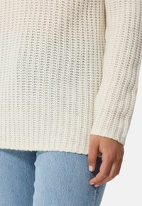 Sweter dzianinowy w kremowym kolorze z teksturowanym, otwartym splotem. Posiada długie rękawy, ściągacze oraz prosty dół, w połączeniu z jasnobłękitnymi dżinsami.