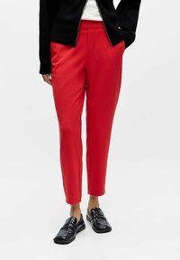 Rote, maßgeschneiderte Hose mit glatter Textur, gestaltet mit Seitentaschen und schmaler Passform, kombiniert mit schwarzen Loafers auf neutralem Hintergrund.