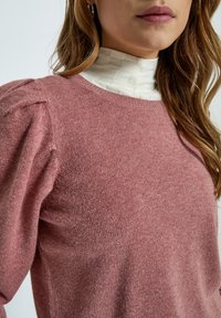 Light pink sweater with a soft texture and balloon sleeves, featuring a crew neckline. Worn over a white ribbed turtleneck.