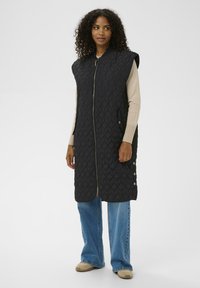 Veste noire matelassée avec fermeture éclair à l'avant, poches latérales et boutons aux épaules, portée par-dessus un haut beige à manches longues et un jean bleu à jambes larges.