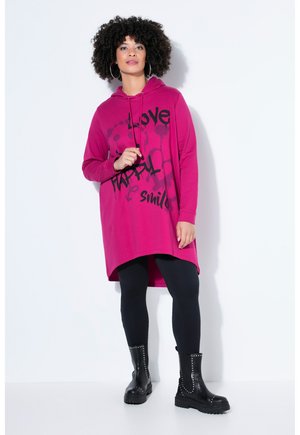 Kvinde med krøllet hår iført store øreringe, en lang lyserød hættetrøje med sort tekst, sorte leggings og sorte støvler med nitter, stående mod en hvid væg.