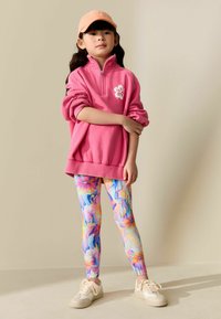 Svetloružová mikina s kvetinovým vzorom, v kombinácii s farebnými legínami s vírivým vzorom a béžovými teniskami. Oranžová čiapka dopĺňa vzhľad.