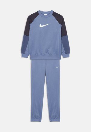 Sininen ja tummansininen Nike-harjoitteluasu, jossa on valkoiset swoosh-logot. Varustettu pitkähihaisella collegepaidalla ja siihen sopivilla housuilla.