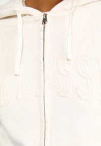 Felpa con zip di colore crema, caratterizzata da lettere bianche in rilievo. Realizzata in un morbido tessuto dalla superficie liscia, include una zip argento e lacci.