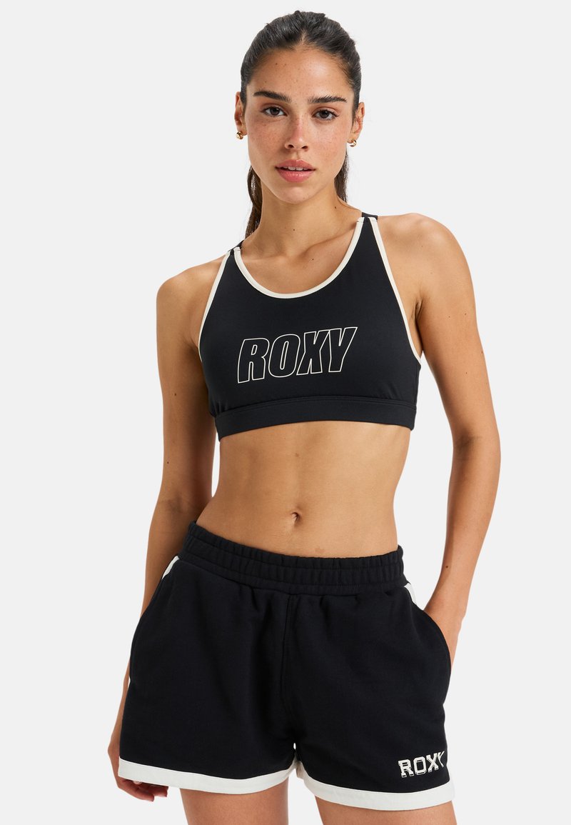 Sujetador deportivo negro con ribete blanco y el logo "ROXY", combinado con pantalones cortos negros con detalles blancos y un ajuste relajado.