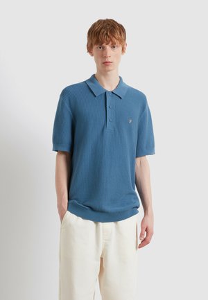 Jong persoon met kort rood haar draagt een gestructureerd blauw poloshirt met korte mouwen en crème wijde broekspijpen, staat met handen in de zakken.