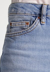 Světle modré džíny s vysokým pasem, vybavené malou přední kapsou, měděnými nýty a viditelnými švy.