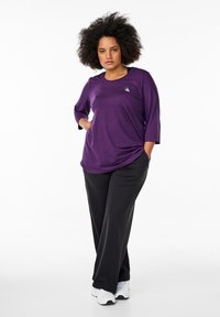 Paarse sporttop met driekwart mouwen en een klein logo op de borst, gecombineerd met zwarte, losse broek en witte sneakers.