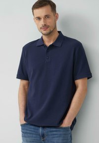Granatowa koszulka polo z krótkim rękawem, z trzydziestoma guzikami, prążkowym kołnierzykiem i gładką fakturą, w zestawie z jasnoniebieskimi dżinsami.