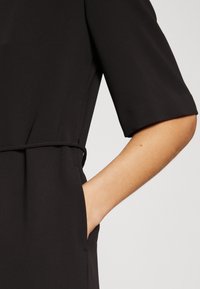 Rochie neagră dintr-un material texturat, cu mâneci scurte, talie ajustată și un detaliu de buzunar lateral. Linii curate și un design minimalist.