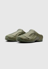Un par de zapatos de color verde oliva, tipo slip-on, con un diseño suave y sin costuras y suelas texturizadas, posicionados uno al lado del otro sobre un fondo gris claro.