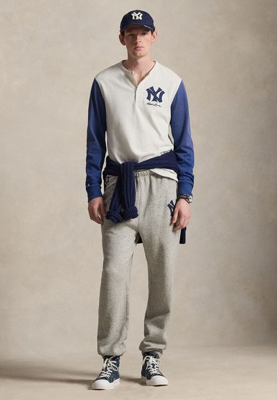 Ung mann med navyblå caps, hvit og blå langermet skjorte, grå joggebukser, navyblå genser knytt rundt livet, og blå høye joggesko.