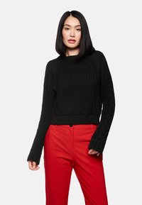 Schwarzer gestrickter Pullover mit gerippter Textur und Zopfmusterdetails. Kürzerer Schnitt mit langen, weiten Ärmeln. Kombiniert mit roten Hosen.