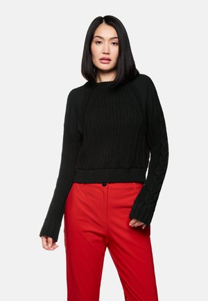 Schwarzer gestrickter Pullover mit gerippter Textur und Zopfmusterdetails. Kürzerer Schnitt mit langen, weiten Ärmeln. Kombiniert mit roten Hosen.