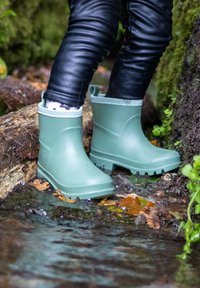 Bottes de pluie en caoutchouc vert avec une surface texturée, à tige haute et bord gris, se tenant dans une eau peu profonde parmi des feuilles et une bûche.