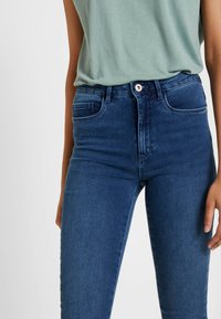 Jeans de ganga azul-escuro com corte justo, cintura alta e desbotamento subtil. Apresenta dois bolsos traseiros e fecho com botão na frente.