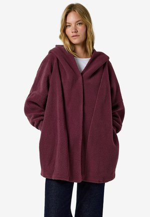 Young woman wearing oversized maroon fleece coat with hood and hands in pockets, standing against white background.