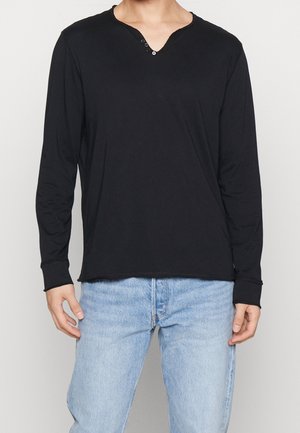 Czarna koszula z długim rękawem i dekoltem w serek, z trzema guzikami, z miękkiego materiału i lekko zaokrąglonym dołem. W połączeniu z jasnoniebieskimi dżinsami.