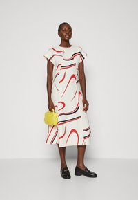 Bíl dress s červenými, námořnickými a černými vlnkovými pruhy, krátké rukávy a tvar A-line. Modelka drží malou žlutou kabelku. Černé mokasíny dokončují vzhled.