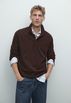 Bruine wollen pullover met een hoge kraag en knoopsluiting, gelaagd over een lichtblauw overhemd. Model draagt blauwe jeans, met handen in de zakken.