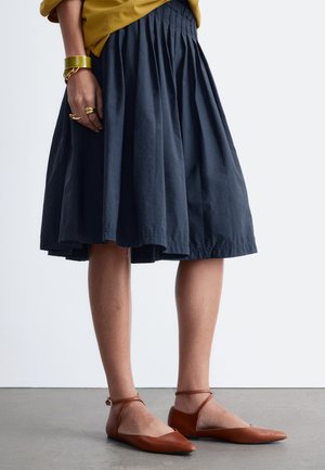 Person iført et mørkeblåt plisseret midi-nederdel, sennepsfarvet top, brune flade sko med ankelrem og gyldne armbånd og ringe på venstre hånd.