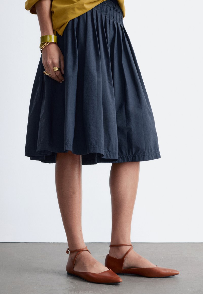 Personne portant une jupe midi plissée bleu marine, un haut moutarde, des ballerines marron à bride à la cheville et des bracelets et bagues dorés à la main gauche.