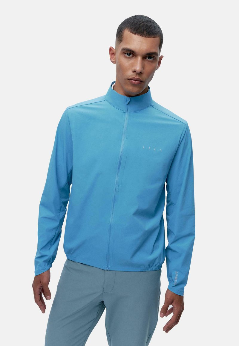 Világoskék, cipzáras dzseki magas gallérral és hosszú ujjakkal, sima, könnyű anyagból készült. Diszkrét logórészlet található rajta.