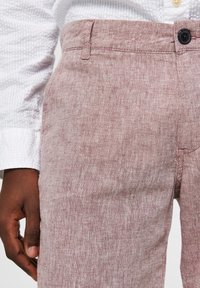 Pantalons en lin rouge clair avec une finition texturée, dotés d'une fermeture à un bouton et de poches latérales, portés avec une chemise blanche texturée.