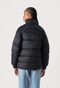 Casaco puffer preto com colarinho alto, apresentando seções acolchoadas e punhos canelados, usado sobre jeans de perna larga de tom azul claro.