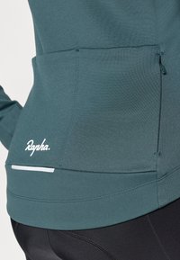 Sudadera de ciclismo en tono verde azulado con un tejido texturizado. Presenta un bolsillo con cremallera, un logo blanco y una franja reflectante en el dobladillo. Diseñada para un ajuste perfecto.