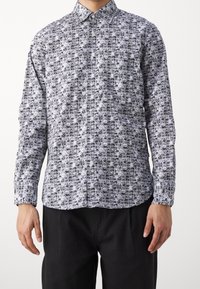 Man som bär en långärmad skjorta med svartvitt kassettbandmönster och svarta byxor, står med armarna avslappnade.