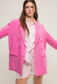 Blazer rosa de gran tamaño con botones de doble botonadura y bolsillos frontales, superpuesto a una camisa blanca con rayas rosas y pantalones cortos anudados.