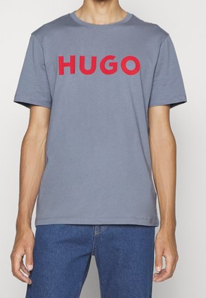 Mężczyzna ubrany w szarą koszulkę z krótkim rękawem z wyraźnym czerwonym napisem "HUGO" na piersi oraz niebieskie dżinsy, stojący na jasnym tle.