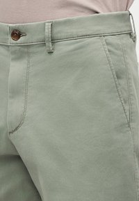 Κοντινό πλάνο ξεθωριασμένου πράσινου παντελονιού με καφέ κουμπί, θηλιές για ζώνη, πλευρική τσέπη και εμφανή ραφές, φορεμένο με μπεζ τοπ.