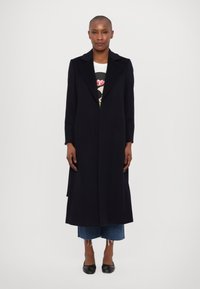 Dlouhý černý kabát s klopovým límcem, strukturovanou látkou a rovný střihem, doplněný grafickým tričkem, zkrácenými modrými džínami a černými balerínami.