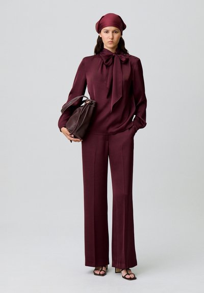 Blusa di seta color bordeaux con fiocco al collo, abbinata a pantaloni a gamba larga dello stesso colore. Tiene una borsa marrone scuro; indossa un foulard abbinato.
