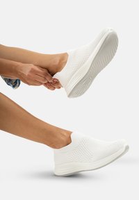 Baskets blanches à enfiler avec un motif en maille, texture lisse et semelle courbée. Présentées aux pieds sur un fond neutre.