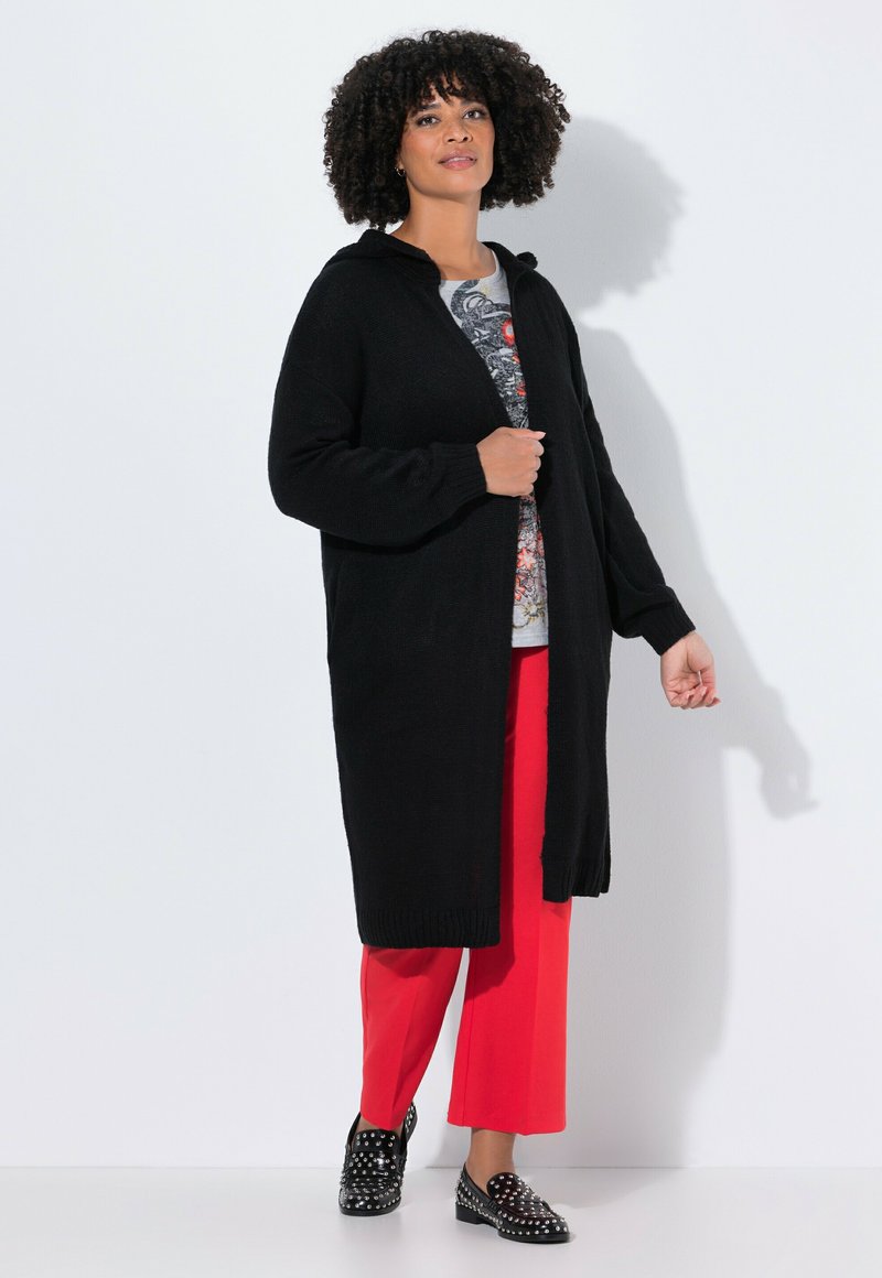 Cardigan noir en tricot avec capuche, à l'avant ouvert, avec des poignets côtelés, porté sur un tee-shirt graphique et associé à un pantalon large rouge et des chaussures studded.
