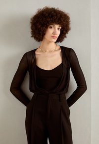 Black sheer long-sleeve top layered over a black fitted tank, paired with high-waisted, textured black trousers. Simple gold necklace accent.